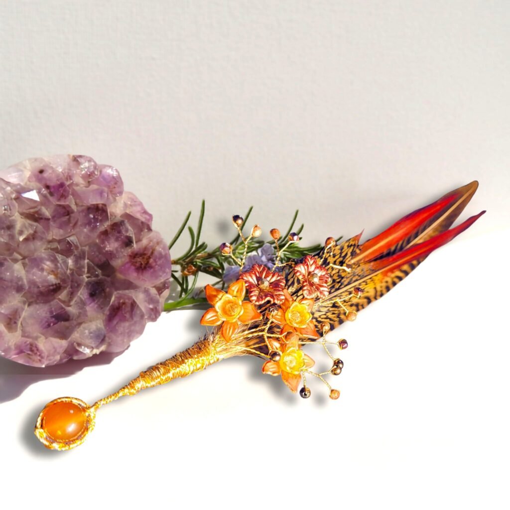 Golden Pheasant feather hatpin in gilt coloured wire with hand painted Lucite flowers and embellished with diamond shaped Magenta Haematite, bronze Haematite and Goldstone rounds with faceted glass crystals and Carnelian at its base.. Designed and crafted by Fiona of Fifi and the Peacock
