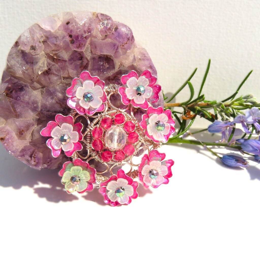 Floral brooch based on a handcrafted wire work filigree style frame with bespoke, hand painted wire work flowers with a Lucite flower and pastel faceted Haematite centre.  The central part of the brooch features a faceted clear Quartz and dyed pink faceted Kyanite.  Designed and crafted by Fiona of Fifi and the Peacock